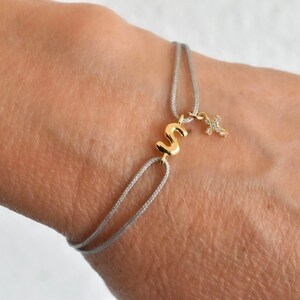May include: A gold-colored initial "S" charm bracelet with a small cross charm. The bracelet features a gray cord and is worn on a wrist. The initial and cross are detailed with small embellishments.