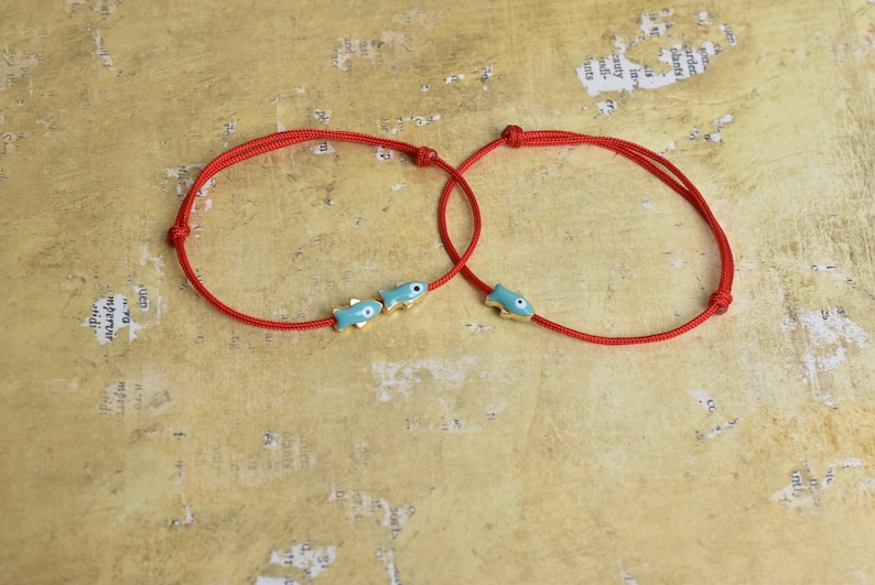 May include: Two red string bracelets, each adorned with small, turquoise fish charms with gold accents. The bracelets are knotted and rest on a textured, tan surface, creating a simple yet charming aesthetic.