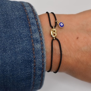 May include: A black cord bracelet with a gold-colored initial charm and a blue and white evil eye charm. The bracelet is worn on a wrist next to a denim jacket.