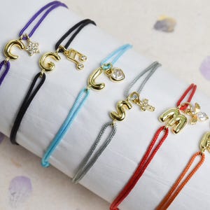 May include: Assortment of colorful string bracelets with gold-colored initial charms and decorative accents. The bracelets feature purple, black, blue, gray, red, and orange cords. Charms include letters, a star, a musical note, a heart, and a cross.