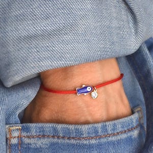 May include: Red string bracelet featuring a blue fish charm with a white eye and a small, clear charm. The bracelet is worn on a wrist, against a denim jacket.