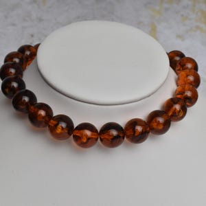 May include: A necklace of large, round amber beads. The beads are a rich, translucent brown color and are strung together to form a choker-style necklace. The necklace is displayed on a white bust.