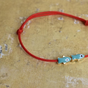 May include: Red cord bracelet featuring two turquoise fish charms with gold accents. The bracelet is knotted and rests on a textured, golden-toned surface. A simple, nautical-themed accessory.