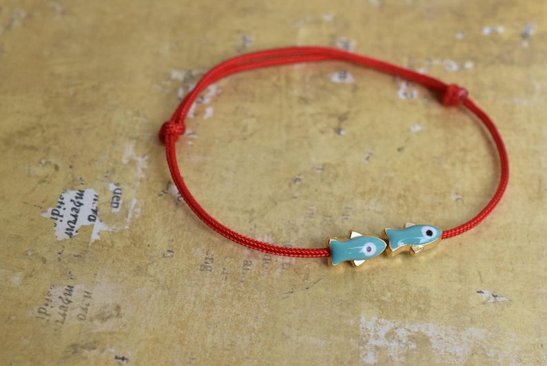 May include: Red cord bracelet featuring two turquoise fish charms with gold accents. The bracelet is knotted and rests on a textured, golden-toned surface. A simple, nautical-themed accessory.