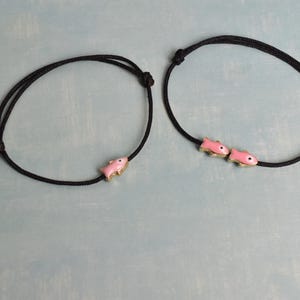 May include: Two black cord bracelets, each featuring a pink fish charm with gold-colored accents. One bracelet has a single fish, the other has two. The bracelets are displayed on a light blue surface.