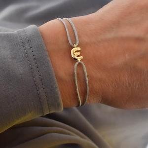 May include: A gold-colored letter "E" charm bracelet with a gray cord. The bracelet is worn on a person's wrist, with the gray cord and gold charm as the focal point. The bracelet is simple and elegant.