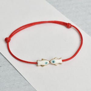 May include: Red string bracelet featuring two small, white fish charms with blue eyes and gold accents. The bracelet is tied with knots and is displayed on a white and gray surface. A simple, elegant piece of jewelry.