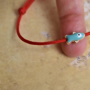 May include: Red string bracelet featuring a turquoise fish charm with gold-colored accents. The fish has a white eye. The bracelet is knotted at the end. The background is a light brown color.