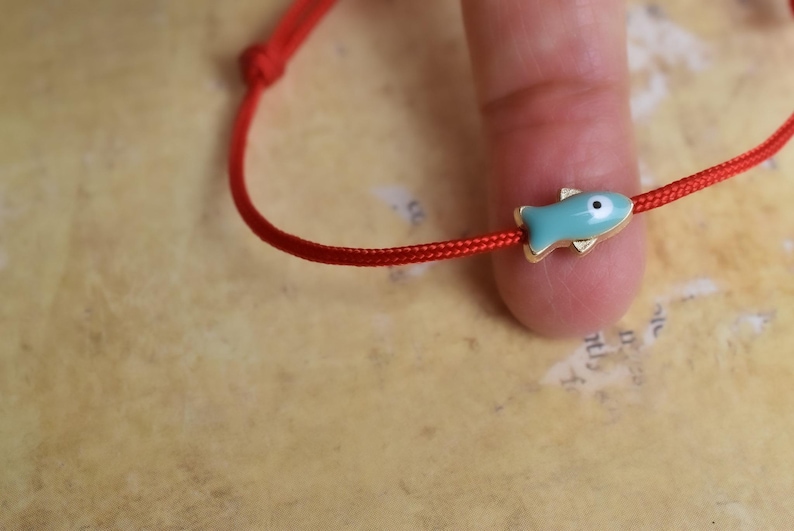 May include: Red string bracelet featuring a turquoise fish charm with gold-colored accents. The fish has a white eye. The bracelet is knotted at the end. The background is a light brown color.