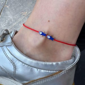 May include: Red string anklet with two blue fish charms with white and black eye details. The anklet is worn on an ankle next to a silver sneaker.