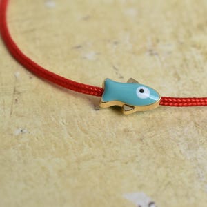 May include: A red cord bracelet with a turquoise fish charm. The fish charm has gold-colored accents and a white eye. The bracelet is displayed on a textured, light-colored surface.