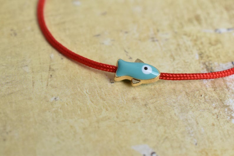 May include: A red cord bracelet with a turquoise fish charm. The fish charm has gold-colored accents and a white eye. The bracelet is displayed on a textured, light-colored surface.