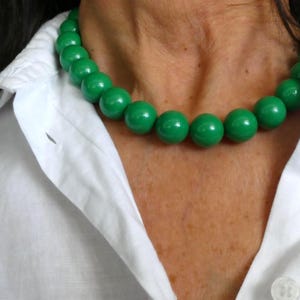 May include: A green beaded necklace with large, round beads. The necklace is worn over a white collared shirt. The beads are a vibrant, glossy green color, creating a bold statement piece.