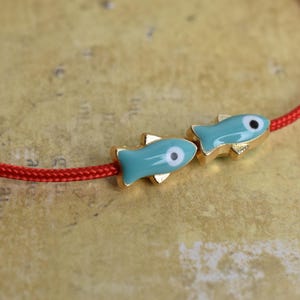 May include: A red cord bracelet featuring two turquoise fish charms with gold-colored accents. The fish have a white and black eye design. The bracelet is set against a textured, tan background.