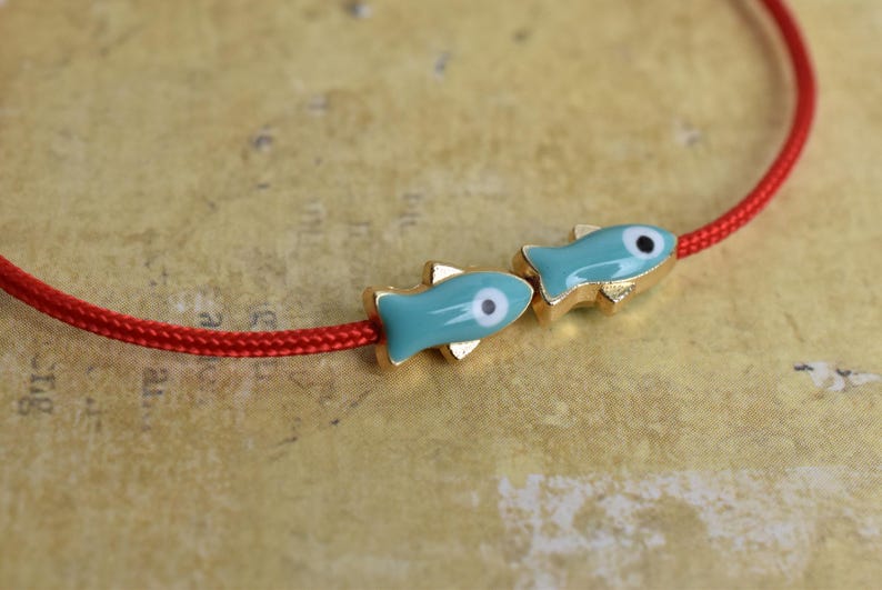 May include: A red cord bracelet featuring two turquoise fish charms with gold-colored accents. The fish have a white and black eye design. The bracelet is set against a textured, tan background.