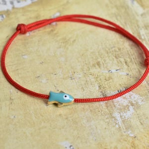 May include: A red cord bracelet featuring a small, light blue fish charm with gold accents. The bracelet is knotted and designed to be adjustable. The fish charm has a white eye. Jewelry.