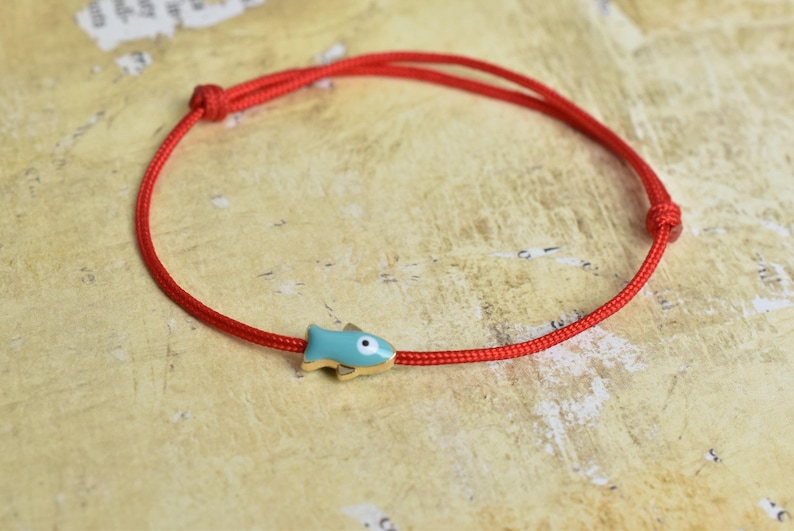 May include: A red cord bracelet featuring a small, light blue fish charm with gold accents. The bracelet is knotted and designed to be adjustable. The fish charm has a white eye. Jewelry.
