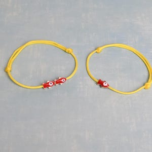 May include: Two yellow cord bracelets with red and white fish-shaped charms. The bracelets are knotted and lie on a light blue surface. The charms have a small eye design.