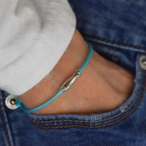 May include: A turquoise cord bracelet with a silver fish charm. The bracelet is worn on a wrist, with the person wearing a white shirt and blue jeans. The jeans have a pocket visible.