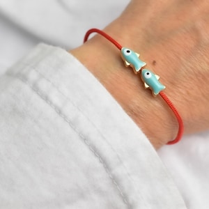 May include: Red string bracelet featuring two light blue fish charms with gold-colored accents. The fish charms have black eye details. The bracelet is worn on a wrist, against a white shirt.