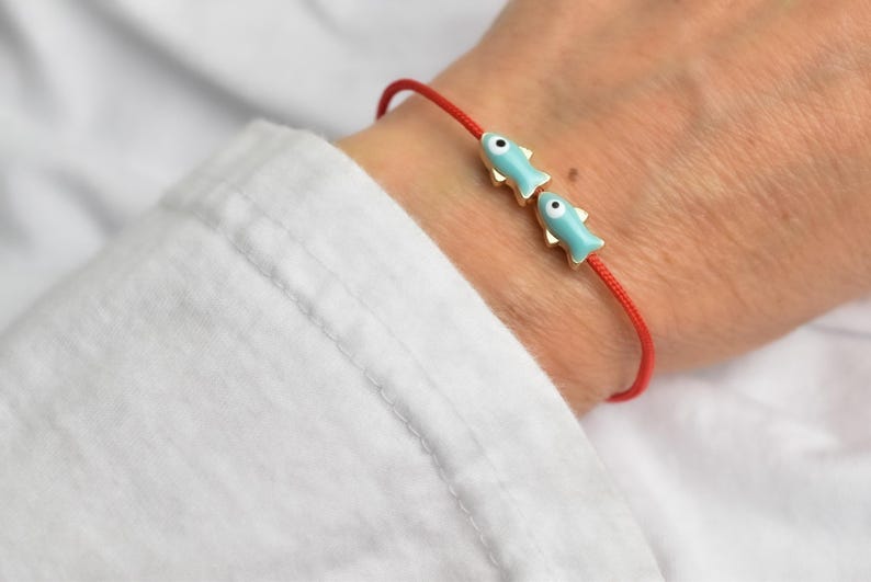 May include: Red string bracelet featuring two light blue fish charms with gold-colored accents. The fish charms have black eye details. The bracelet is worn on a wrist, against a white shirt.