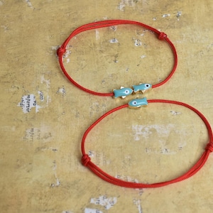 May include: Two red cord bracelets. One bracelet features three small, light blue fish charms with gold accents. The other bracelet is a simple red cord. The bracelets are displayed on a textured, tan surface.