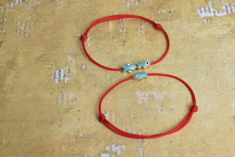 May include: Two red cord bracelets. One bracelet features three small, light blue fish charms with gold accents. The other bracelet is a simple red cord. The bracelets are displayed on a textured, tan surface.