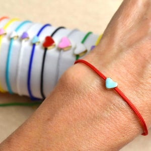 May include: A red cord bracelet with a light blue heart charm. Other bracelets with different colored cords and heart charms are visible in the background. The heart charms have a gold border.