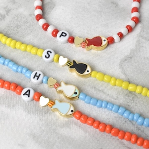 May include: A collection of beaded bracelets in red, white, yellow, blue, and orange. Each bracelet features a gold-colored fish charm and letter beads spelling out names. The bracelets are on a white surface.