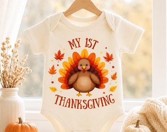 May include: A cream-colored short-sleeve baby bodysuit featuring a central graphic of a brown turkey with a colorful orange, yellow, and red tail. The text "MY 1ST THANKSGIVING" is printed in brown above and below the turkey, surrounded by autumn leaves.