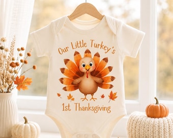 May include: A cream-colored short-sleeve baby bodysuit featuring a central graphic of a cartoon turkey with orange and brown feathers. The text "Our Little Turkey's 1st Thanksgiving" is printed in brown above and below the turkey illustration.