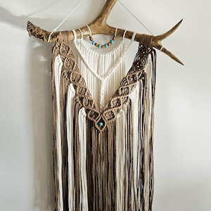 May include: A macrame wall hanging, approximately 40 inches long and 19 inches wide, featuring a natural antler base. The design incorporates brown and white knotted cords, accented with turquoise beads, creating a bohemian aesthetic.