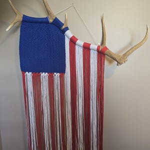 May include: A wall hanging in the style of the American flag, with a blue woven section and red and white fringe. The artwork is mounted on deer antlers, adding a rustic touch. The piece is designed for wall display.