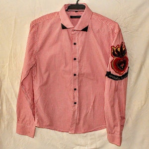 May include: Red and white gingham long-sleeved button-down shirt with a black collar and a black heart-shaped patch on the right sleeve. The patch has gold embroidery and the text "Amor Vincit Omnia".