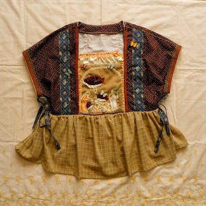 Op de afbeelding: Een bruin en beige patchwork top met een gerimpelde rok en strik sluitingen. De top heeft een centraal paneel met een geborduurde scène van paddenstoelen en figuren.