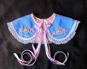 Può includere: Un colletto in tessuto blu con bordino in pizzo bianco e lacci in nastro rosa. Il colletto è ricamato con fiori e presenta un motivo floreale rosa e bianco sul retro.