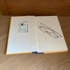 Puede incluir: Un libro abierto con una cubierta amarilla y un lomo azul. Una pequeña pegatina con el texto "from the library of" está en la portada. Un par de gafas con montura plateada está sobre las páginas abiertas.