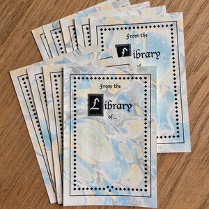 Puede incluir: Un conjunto de ocho tarjetas de ex libris en blanco con un fondo marmoleado azul y amarillo. Las tarjetas tienen un borde negro con un patrón de diamantes y el texto "From the Library of..." en letra cursiva negra.