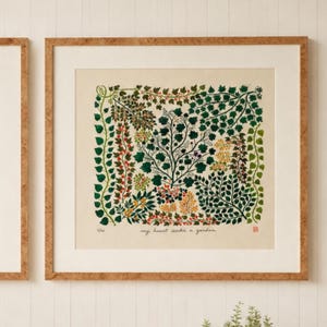 以下が含まれることがあります： クリーム色の背景に、様々な緑の葉、蔓、小さな果実が描かれた詳細な植物画の正方形の額入りアートプリント。下部には黒字で「my heart seeks a garden」というテキストと、小さな赤いスタンプが押されています。