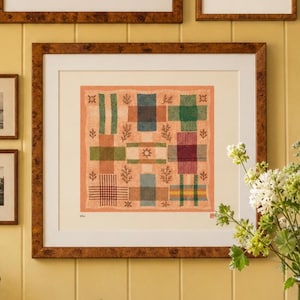 May include: A square framed print featuring a grid of nine rectangular blocks in various colours including green, blue, purple, and brown, set against a peach background with small leaf motifs and a central star symbol, enclosed in a burl wood frame.