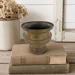 May include: A tarnished brass bowl with a wide, flared rim and a pedestal base. The exterior surface features a horizontal band of embossed leaf patterns. The bowl is positioned on top of two stacked, cloth-bound hardcover books.