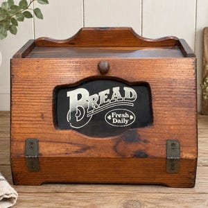 May include: A brown wooden bread box with a hinged front door featuring a central black panel. The panel displays the white text BREAD and Fresh Daily. The box includes a small wooden knob for opening and two metal hinges at the bottom base.