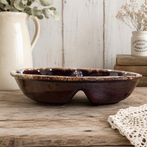 May include: A dark brown ceramic serving dish with a divided interior and a distinct arched cutout at the base. The rim features a textured, lighter brown drip glaze pattern that contrasts with the smooth, glossy finish of the main body.