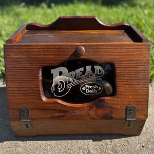 May include: A rectangular wooden bread box with a dark brown finish, featuring a hinged front door with a central glass panel. The glass displays the white text "BREAD" and "Fresh Daily" with a small clover icon. It has two metal hinges.