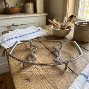 May include: A silver-toned metal trivet featuring a geometric, elongated frame with two central wavy bars and four short legs. The structure is constructed from smooth, polished metal with textured, ribbed detailing along the outer side handles.