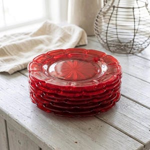 May include: A stack of five circular red glass plates featuring a scalloped edge design and a central starburst pattern. The plates are translucent and have a smooth, reflective surface, showcasing a consistent deep red color throughout the set.