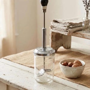 May include: A clear glass jar with embossed measurement markings, featuring a silver-toned metal lid and a vertical plunger mechanism topped with a round black handle. The device is designed for mixing or churning, with a metal agitator inside.