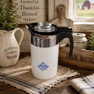 May include: A white percolator coffee pot with a black handle and lid, featuring a clear glass knob on top and a blue floral graphic on the front. The body is cylindrical with a metallic silver band near the top rim, contrasting with the white base.
