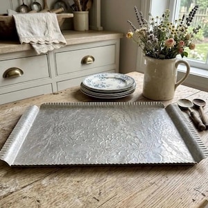 May include: A rectangular silver-toned metal serving tray featuring an embossed floral and grape vine pattern across its surface. The tray has raised, scalloped edges on the sides and a textured finish, providing a decorative base for serving items.