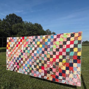 May include: A rectangular patchwork quilt composed of numerous small, multi-colored fabric squares in various patterns, including solids, florals, and plaids. The squares are joined with small red fabric ties at each corner intersection.